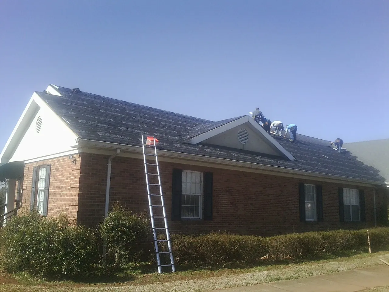 Crew tearing shingles off a brick ranch roof with a ladder set — Metal Roof Repair work by our crew in Ault