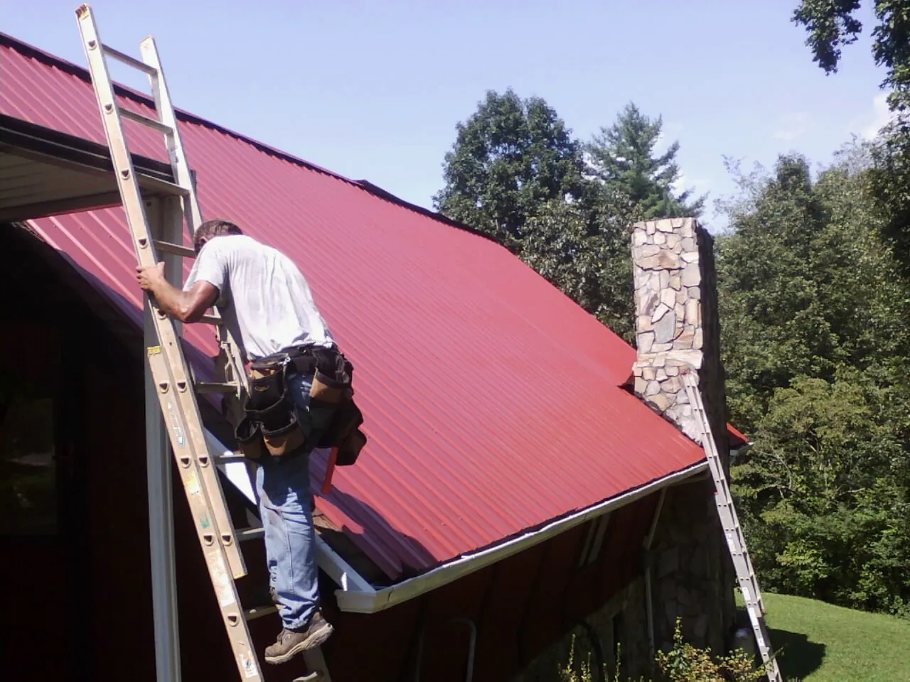 Roofer on a ladder beside a red metal roof and stone chimney — Soffit & Fascia Repair by Roofing Brothers in Ault